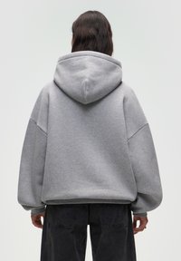 Felpa grigia oversize realizzata in tessuto morbido, con un grande cappuccio e cuciture delle spalle scese. Presenta polsini e orlo a costine, abbinata a pantaloni neri.