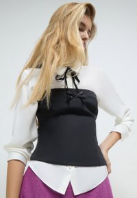 Top bianco e nero caratterizzato da un design aderente con un papillon e dettagli in perle, completato da maniche lunghe e voluminose e uno stile di stoffa stratificato.