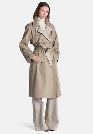 Femme portant un trench-coat beige ceinturé par-dessus un col roulé blanc et un pantalon à jambes larges, debout les mains dans les poches devant un fond uni.