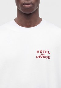 Gros plan sur une personne portant une chemise blanche avec un texte rouge "HÔTEL DU RIVAGE" sur la poitrine gauche.