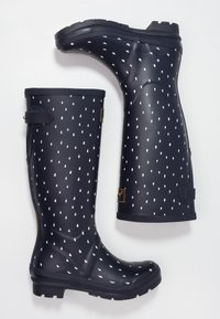 Une paire de bottes de pluie en caoutchouc noires avec un motif de gouttes de pluie blanches, une sangle réglable près du haut et des semelles épaisses à crampons.