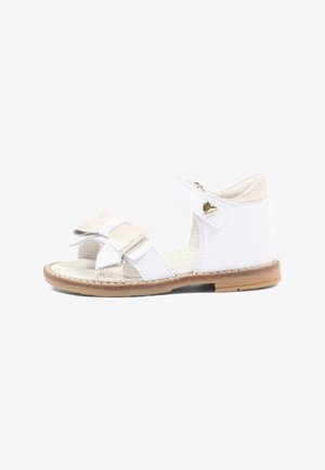 Biała sandał dziecięcy z paskiem wokół kostki, złotym serduszkiem, kokardką na przednich paskach oraz jasnobrązową podeszwą, pokazany z boku na białym tle.