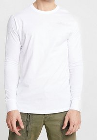 Vit långärmad t-shirt i bomull, med rund halsringning och subtil branding på bröstet. Slät textur med tighta ärmar.