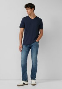 Granatowy t-shirt z dekoltem w serek i krótkimi rękawami, w połączeniu z jasnoniebieskimi dżinsami. Model nosi białe sneakersy z szarymi akcentami. Prosty design i fason.