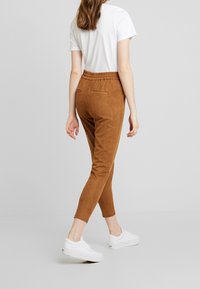Pantalones elásticos de color marrón claro con pierna ajustada, cintura elástica y bolsillos traseros, combinados con zapatillas deportivas blancas y una camiseta blanca de manga corta.