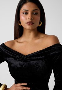 Kobieta o prostych, ciemnych włosach nosi czarną, aksamitną bluzkę z odkrytymi ramionami, duże złote kolczyki w kształcie koła oraz grubą złotą bransoletkę, na jednolitym tle.