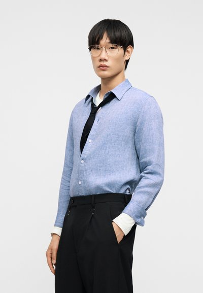 Άνδρας που φοράει γαλάζιο πουκάμισο με κουμπιά πάνω από λευκή μπλούζα με μακριά μανίκια, χαλαρή μαύρη γραβάτα, μαύρο παντελόνι και γυαλιά, στέκεται με το χέρι στην τσέπη.