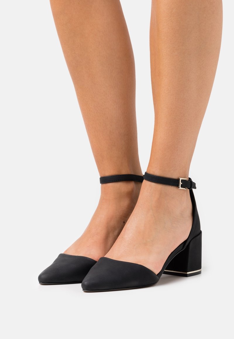 Black leather heels with a pointed toe and ankle strap. Block heel with a gold accent and smooth texture.
