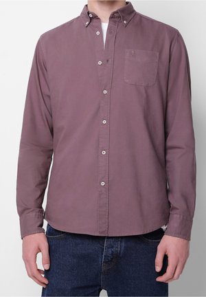 Mann trägt ein mauvefarbenes, langärmeliges Hemd mit Knöpfen und Brusttasche sowie blaue Jeans vor einem schlichten Hintergrund.