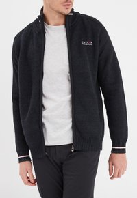 Schwarze gestrickte Zip-Up-Kardigan mit hohem Kragen, strukturiertem Stoff, gerippten Bündchen und dezenten gestreiften Details. Getragen über einem hellgrauen Hemd.