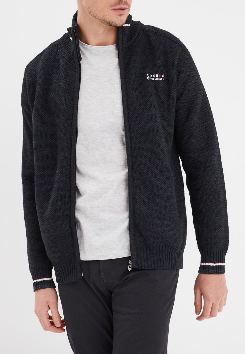 Schwarze gestrickte Zip-Up-Kardigan mit hohem Kragen, strukturiertem Stoff, gerippten Bündchen und dezenten gestreiften Details. Getragen über einem hellgrauen Hemd.