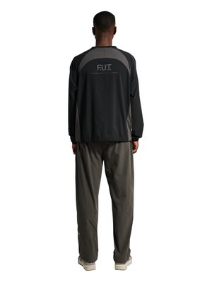 Mann, der mit dem Rücken zur Kamera steht, trägt ein schwarz-graues Langarmoberteil mit der Aufschrift „F.U.T. Physical Education & Training“ auf dem Rücken und lockere dunkle Hose.