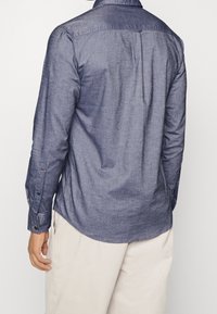 Camisa de manga larga azul oscuro hecha de tela texturizada, con cuello abotonado, detalle de canesú trasero y dobladillo redondeado.