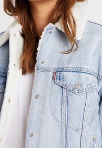 Jasnoniebieska kurtka dżinsowa z białym kołnierzem wyłożonym futerkiem sherpa, srebrnymi elementami metalowymi i kieszenią na piersi; cechuje ją widoczne przeszycie i wszywka z logo.