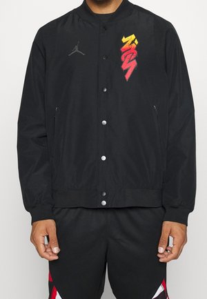 Man wearing black buttoned jacket with gray Jumpman logo on left chest and red-yellow stylized graphic on right chest, paired with black and red shorts.