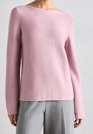 Woman wearing a light pink long-sleeve knit top paired with high-waisted gray trousers against a plain background.