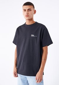 T-shirt in cotone nero con maniche corte, caratterizzata da un piccolo logo bianco nella parte superiore sinistra. Vestibilità casual, texture liscia, abbinata a jeans azzurri chiari.