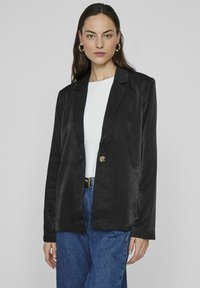 Juodas švarkas, pagamintas iš lygaus audinio, su vienu auksiniu mygtuku, notch tipo apykakle ir ilgomis rankovėmis, derinamas su baltu palaidiniu ir mėlynomis džinsomis.