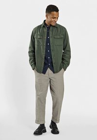 Green button-up jacket with two chest pockets over a dark blue shirt, paired with light beige trousers and black shoes. Smooth textures.