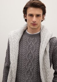 Maglione grigio lavorato a maglia con un motivo a treccia testurizzato, completato da una sciarpa a scialle bianca drappeggiata sulle spalle. Aspetto strutturato e a strati.