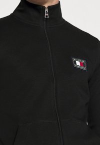 Felpa nera con zip e colletto alto, caratterizzata da una patch logo rettangolare in blu, bianco e rosso con la scritta "HILFIGER". Tessuto morbido, design aderente.