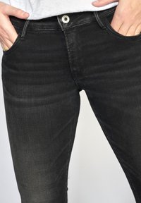Jeans noirs ajustés avec une texture délavée, design à cinq poches et fermeture à bouton en argent. Tissu lisse avec un léger stretch.