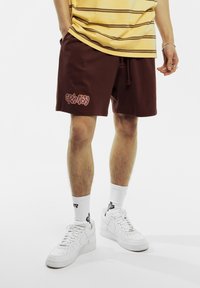 Bruine shorts met een trekkoord, met het "GRIMEY" logo in oranje. Gepaard met witte Nike sneakers en een gestreept geel shirt. Textuurstof.