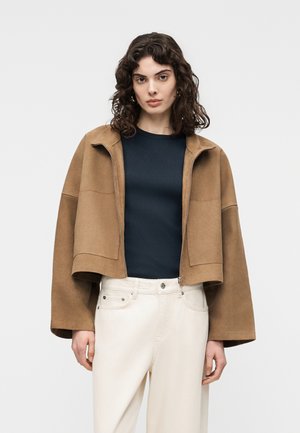 Kvinde med krøllet mørkt hår iført en cropped brun jakke over en sort top og hvide højtaljede bukser, stående foran en ensfarvet baggrund.