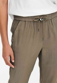Gros plan d'une personne portant un pantalon à cordon de serrage vert olive et une chemise blanche, la main reposant près de la poche gauche.