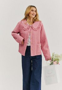 Růžová prošívaná bunda s velkým volánovým límcem, zapínáním na knoflíky a náprsní kapsou; nošena přes bílý top a doplněna tmavě modrými kalhotami.