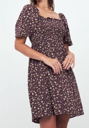 Mujer con vestido morado de flores, mangas abullonadas y corpiño fruncido, manos relajadas cerca de la cintura, fondo blanco.