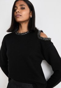 Μαύρο πουλόβερ με υφασμάτινη, διακοσμημένη λαιμόκοψη και ανοίγματα στους ώμους. Διαθέτει μαλακό πλεκτό υλικό και χαλαρή εφαρμογή.