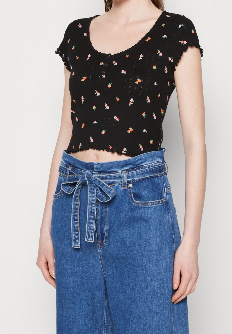 Černý krátký top s barevným květinovým vyšíváním, krátkými rukávy a vlnitým okrajem. Nosený s modrými džíny s vysokým pasem a zavázaným opaskem.