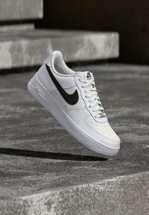 Witte Nike Air Force 1 sneaker met zwarte swoosh, veters gestrikt, zwevend in de lucht boven een gestructureerd betonnen oppervlak buiten.
