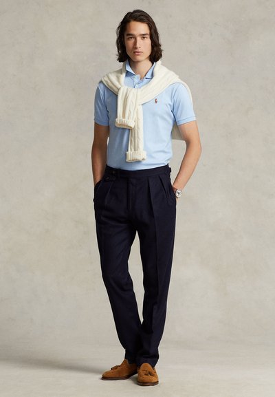 Nuori mies, jolla on tummat hiukset, pukeutunut vaaleansiniseen poolopaitaan, valkoinen villapaita olalla, tummat laskostetut housut ja ruskeat loaferit, seisomassa.