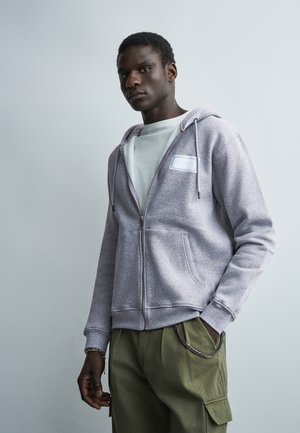 Mladý muž majúci na sebe svetlosivú mikinu na zips cez biele tričko a olivovo zelené cargo nohavice, stojaci pred jednoduchým svetlým pozadím.