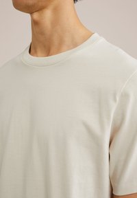 Tricou din bumbac bej deschis, cu decolteu rotund și mâneci scurte. Textură netedă, cu cusături minime și o croială lejeră.