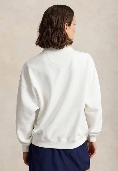Femme aux cheveux courts marron, vue de dos, portant un sweat-shirt blanc ample et une jupe bleu foncé sur un fond beige uni.
