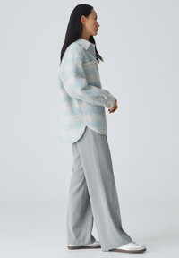 Chemise rayée surdimensionnée en bleu clair et blanc, en tissu texturé, avec un col et une poche ; associée à un pantalon large gris et des baskets blanches.