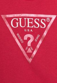 Röd bomulls-T-shirt med en triangulär grafik som har "GUESS", ett frågetecken och texten "WASHED JEANS" i vitt och metallisk rosa.
