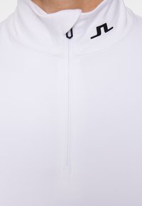 Vit tröja med hög krage och dragkedja fram. Har en liten svart logga på vänster sida nära kragen. Slät textur.