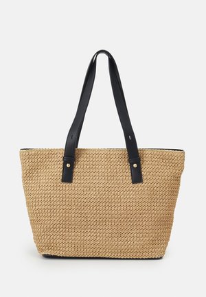 Sac fourre-tout tissé beige avec des bretelles en cuir noir et de petits rivets dorés sur fond blanc.