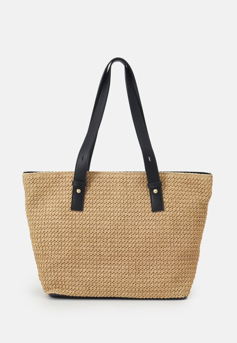 Sac fourre-tout tissé beige avec des bretelles en cuir noir et de petits rivets dorés sur fond blanc.