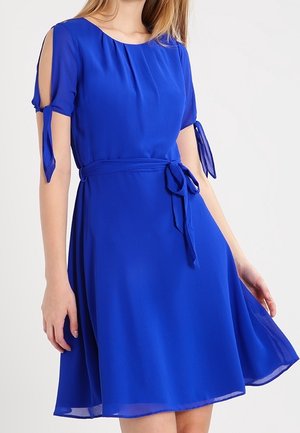 Vrouw draagt een koningsblauwe jurk tot op de knie met korte cold-shoulder mouwen en een tailleband, staand tegen een effen achtergrond.