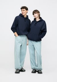 Laivo mėlynos kedės su kiškio kišenėmis, derinamos su šviesiai mėlynomis plačiomis kelnėmis. Abu dėvi juodus sportinius batelius. Paprastas, kasdienis dizainas.