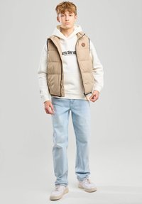 Beige puffer-vesti, jossa on vetoketju, on kerroksittain valkoisen hupparin päällä, jossa on mustaa tekstiä. Vaaleansiniset farkut ja valkoiset lenkkarit täydentävät asukokonaisuuden.