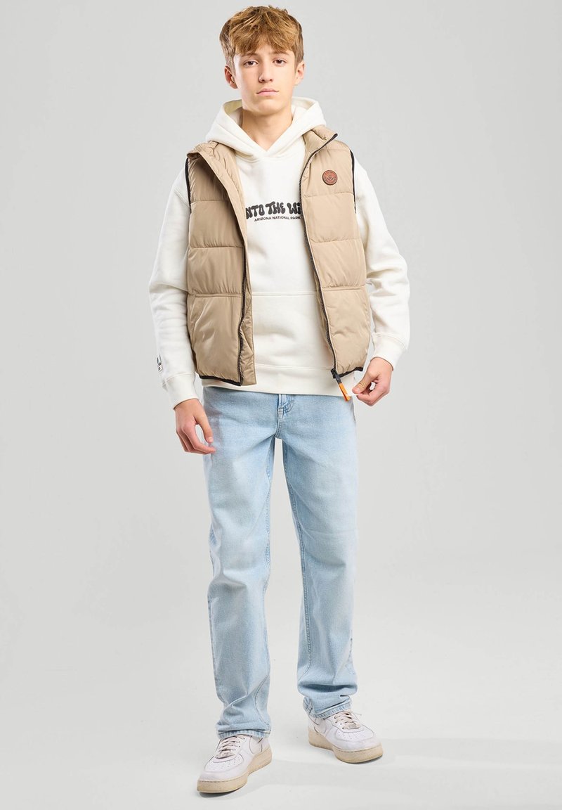 Beige puffer-vesti, jossa on vetoketju, on kerroksittain valkoisen hupparin päällä, jossa on mustaa tekstiä. Vaaleansiniset farkut ja valkoiset lenkkarit täydentävät asukokonaisuuden.