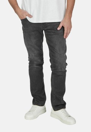 Man draagt donkergrijze spijkerbroek, witte sneakers en een wit overhemd, met één hand in de zak en de andere rustend op de dij.