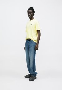 T-shirt gialla in cotone, vestibilità comoda, abbinata a jeans in denim blu, cintura nera e sneakers scure. Design semplice, colori uniformi.