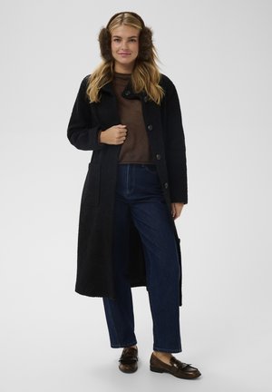 Dolgi črni zunanji plašč z teksturirano tkanino, zapenjanje na gumbe, stranski žepi. Pod njim rjav pulover, modre široke hlače in rjavi loaferji.